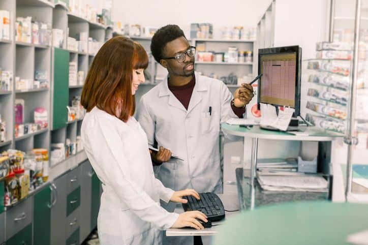 A pharmacist, clinic admin, or health-center leader working side-by-side with a technical specialist (or simply two professionals) looking at a dashboard, report, or pricing data on a laptop or desktop monitor.